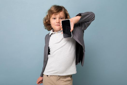 Handsome positive boy with curly hair wearing suit holding phone isolated over blue background looking at camera and showing smartphone with empty display screen. Cutout