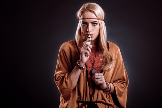 Young woman in the Boho style blowing smoke. The blonde on a dark background. Young woman looking into the camera