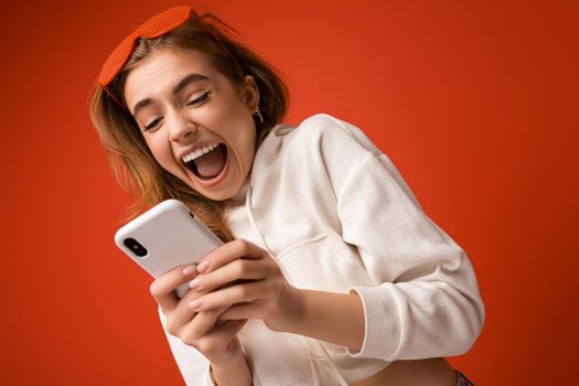 Beautiful surprised emotional young blonde woman wearing stylish white hoodie and funny colorful glasses standing isolated over colourful background holding and using mobile phone communicating online on the internet looking at gadjet and shouting.