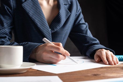 Corporate consultant analyze financial diagram. Close up woman hand with pen and coffee cup on wooden desk. Analyst in suit working at workplace in office. Business accounting and management.