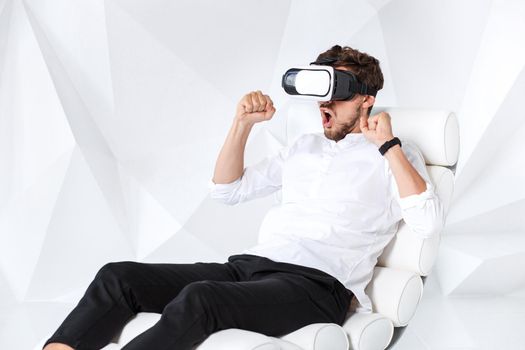 Excited young man is getting experience using VR-headset glasses of virtual reality gesticulating with his hands. A young man sits on a comfortable armchair in a room with white walls