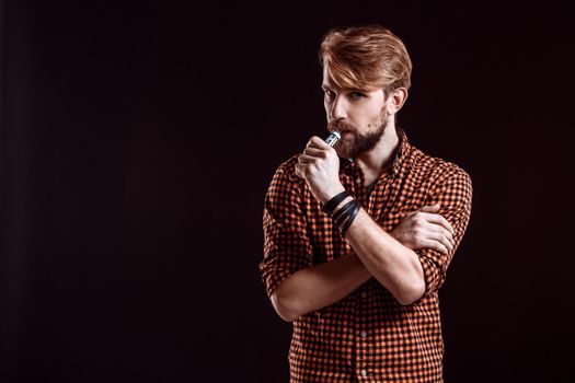 young man wearing a plaid shirt smokes an electronic cigarette on a black background. He looks into the camera