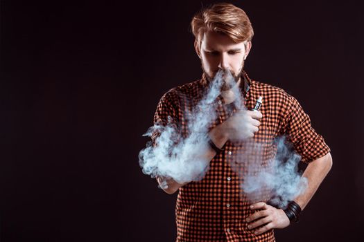 young man wearing a plaid shirt smokes an electronic cigarette on a black background