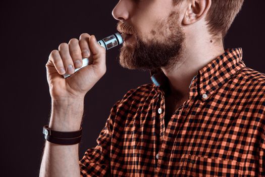 young man wearing a plaid shirt smokes an electronic cigarette on a black background. close-up