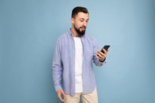 handsome young brunette unshaven man with beard wearing stylish white t-shirt and blue shirt isolated over blue background with empty space holding in hand and using phone messaging sms looking at device screen.