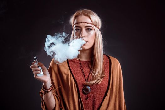 Young woman in the Boho style blowing smoke. The blonde on a dark background. Young woman looking into the camera