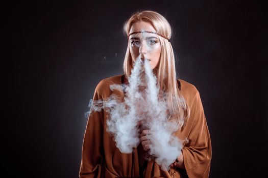 Young woman in the Boho style blowing smoke. The blonde on a dark background. Young woman looking into the camera
