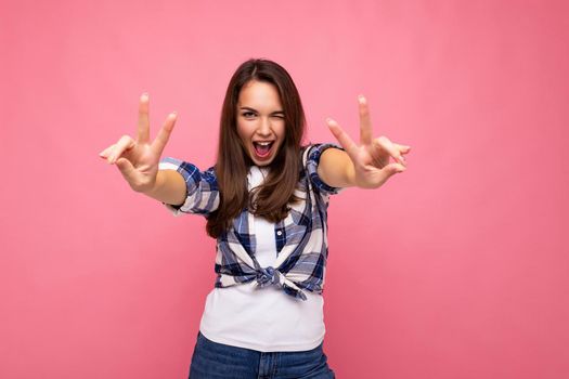 Photo shot of young positive delightful smiling pretty brunette woman with sincere emotions wearing hipster check shirt standing isolated on pink background with free space and showing peace gesture.