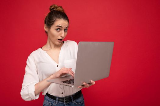 Side profile of Beautiful surprised brunet young woman holding netbook computer looking at camera wearing white shirt typing on keyboard isolated on red background.