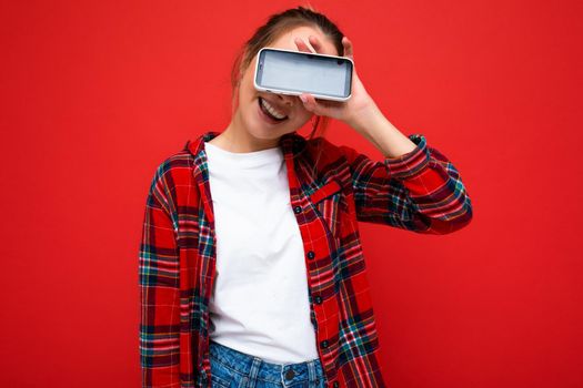Photo of beautiful cute positive young blonde woman standing isolated over red wall wearing casual red shirt and white t-shirt showing mobile phone with empty screen for mockup.
