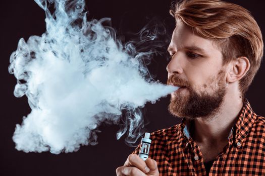 young man wearing a plaid shirt smokes an electronic cigarette on a black background. close-up