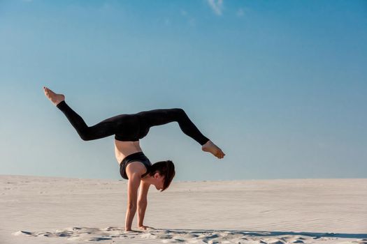 Young woman practicing inversion balancing yoga pose handstand on beach with white sand and bright blue sky