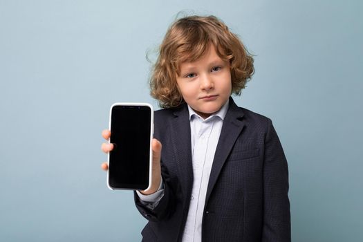 Closeup portrait of Handsome positive boy with curly hair wearing suit holding phone isolated over blue background looking at camera and showing smartphone with empty display screen.