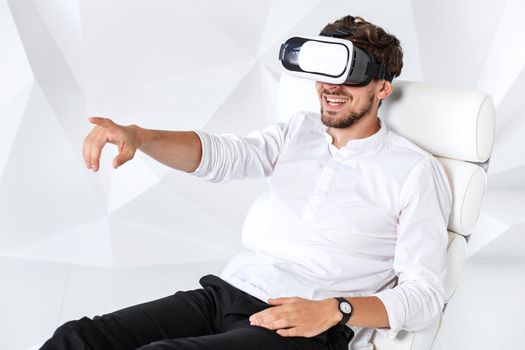 Excited young man is getting experience using VR-headset glasses of virtual reality gesticulating with his hands. A young man sits on a comfortable armchair in a room with white walls