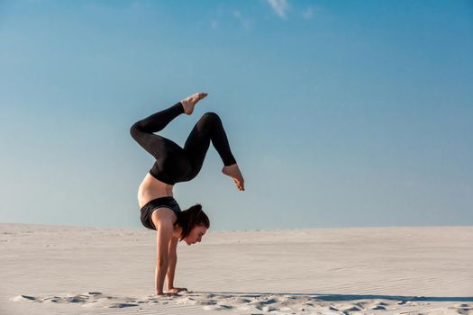 Young woman practicing inversion balancing yoga pose handstand on beach with white sand and bright blue sky