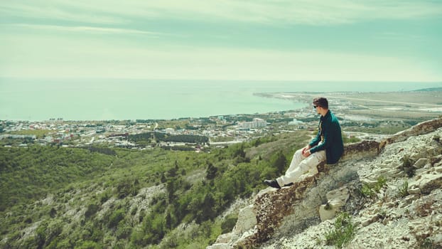 Young man sitting on peak of mountain and enjoying view of sea in summer