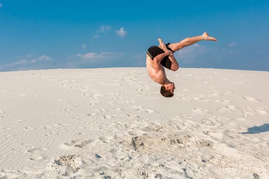 Portrait of young parkour man doing flip or somersault on the sand. Freezed moment of beginning doing bounce.