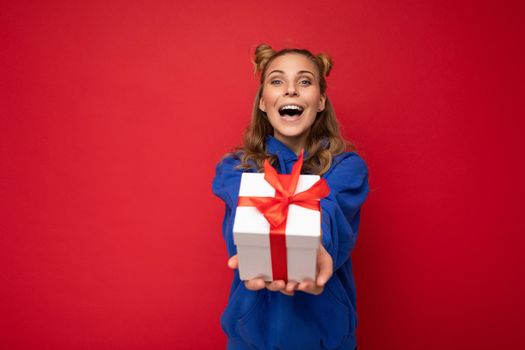 Photo shot of pretty positive surprised amazed young blond woman isolated over colourful background wall wearing trendy outfit look holding gift box and looking at camera.