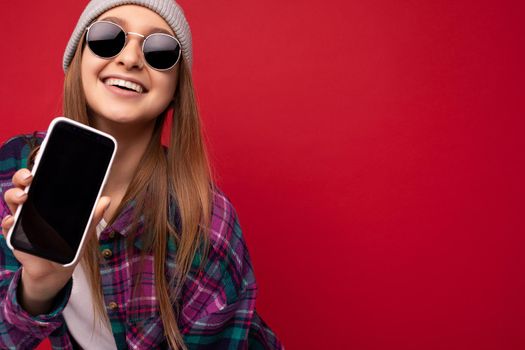 Closeup photo of attractive smiling positive young blonde woman wearing stylish purple shirt and casual white t-shirt grey hat and sunglasses isolated over red background holding in hand and showing mobile phone with empty screen for cutout looking at camera. Copy space
