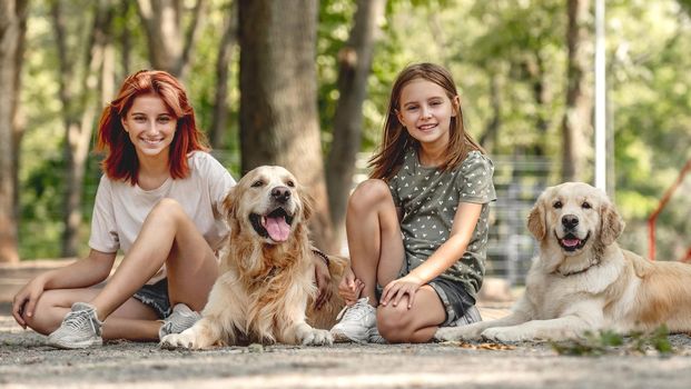 Girls with golden retriever dogs sitting in the park. Sisters with doggy pets outdoors looking at camera