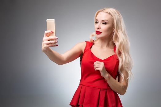 Portrait of a Beautiful successful blonde doing selfie in a red dress on a light background