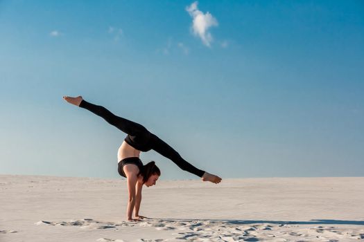 Young woman practicing inversion balancing yoga pose handstand on beach with white sand and bright blue sky