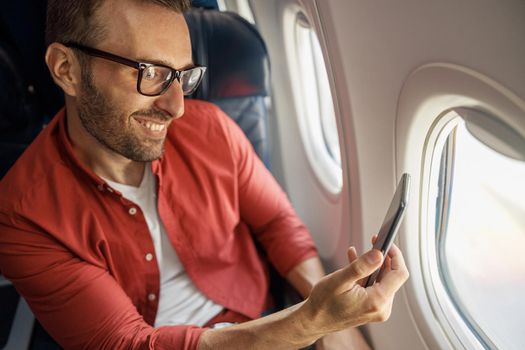 Cheerful caucasian man in casual wear and glasses traveling and taking picture of view outside through airplane window. Technology, travel, vacation, transportation concept
