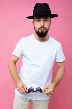 Vertical photo shot of confident brutal good looking young brunet bearded man wearing casual white t-shirt for mockup and stylish black hat poising isolated on pink background with empty space for text and holding sunglasses.