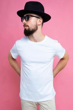 Vertical photo shot of confident brutal good looking young brunet bearded man wearing casual white t-shirt for mockup , sunglasses and stylish black hat poising isolated on pink background with empty space for text.