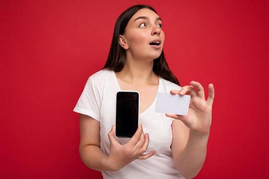 Photo of beautiful surprised young brunette woman wearing white t-shirt isolated on red background holding credit card and mobile phone with empty display for cutout looking to the side.