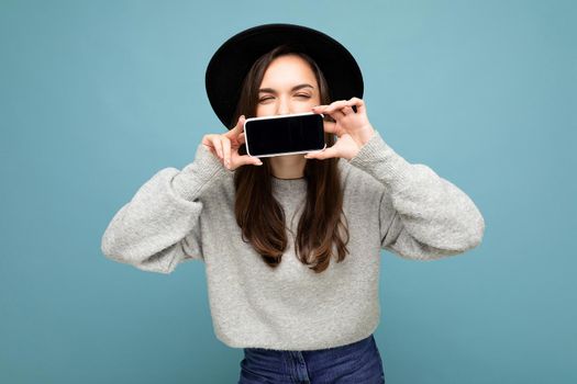 Photo of Beautiful positive woman person wearing black hat and grey sweater holding mobilephone showing smartphone isolated on background with close eyes.Mock up, cutout, free space