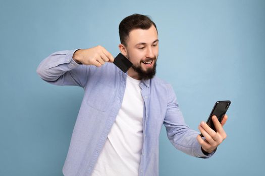 Handsome man wearing everyday clothes isolated on background wall holding and using phone and credit card making payment looking at smartphone screen,