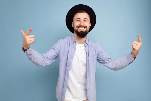 Photo shot of positive happy smiling good looking young brunet bearded man wearing casual blue shirt and white t-shirt and stylish black hat poising isolated on blue background with free space for text.