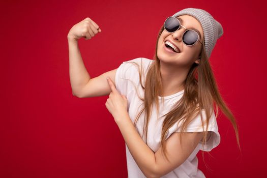 Portrait photo of young emotional positive happy beautiful attractive dark blonde woman with sincere emotions wearing casual white t-shirt with empty space for mockup, grey hat and sunglasses isolated over red background with copy space and showing hand strength.