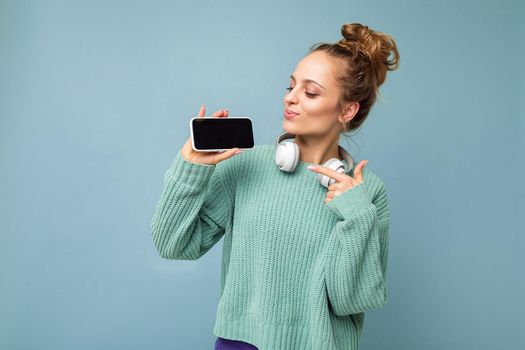 Attractive happy smiling young woman wearing stylish casual outfit isolated on background wall holding and showing mobile phone with empty display for mockup wearing white bluetooth headphones listening to music looking and pointing finger at gadjet.