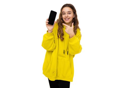 Photo of a beautiful adorable smiling positive funny brunette teenage girl in a stylish yellow sports hoodie holding a mobile phone with a black blank screen isolated on a white background with free space for text.