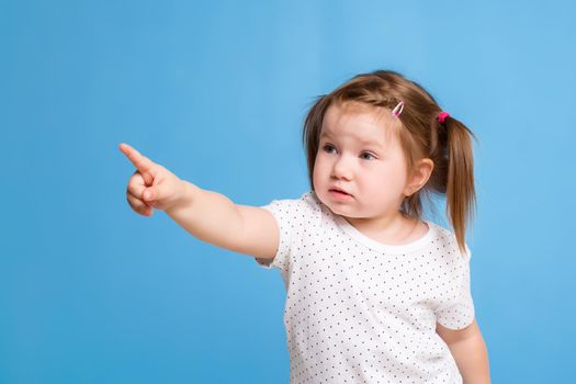 Funny kid in white T-shirt on blue background. Little pretty girl Shows a finger. Copy space for text. Sale, holidays, birthday party concept.