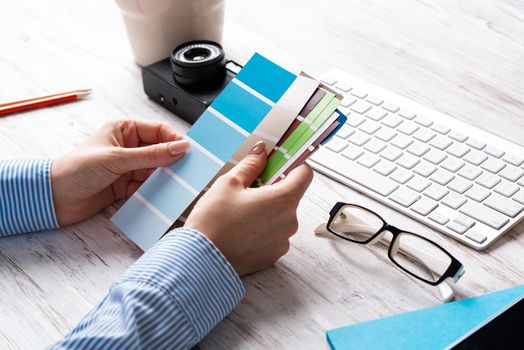 Interior designer holding color swatches at wooden desk. Office workplace with computer keyboard and camera. Brainstorming and creativity. Selection of color palette for web or product design.