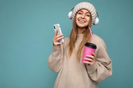 Attractive smiling young blonde woman wearing beige sweater and beige hat white headphones isolated over blue background holding in hand and using mobile phone drinking beverage and listening to music looking at camera and enjoying. empty space