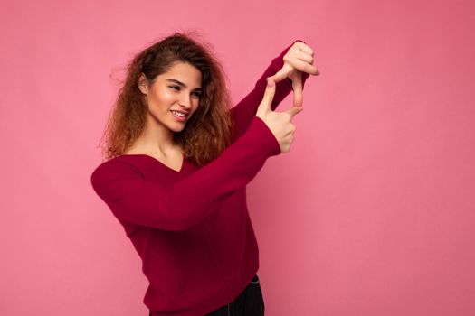 Portrait of young positive pretty brunette curly woman with sincere emotions wearing trendy pink pullover isolated on pink background with copy space and showing camera gesture with hands taking photo.