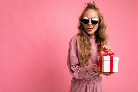 Photo shot of young beautiful attractive cute positive happy curly blonde woman wearing trendy and stylish pink sport suit and sunglasses isolated over pink background with copy space for text. Holding white gift box with red ribbon. Birthday concept.