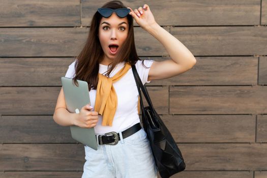 Photo of attractive beautiful shocked amazed surprised charming young brunette girl wearing stylish clothes holding computer laptop and black sunglasses in white t-shirt and light blue jeans in the street near brown wall.
