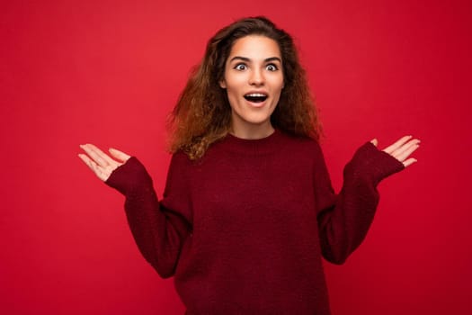 Young beautiful curly brunette woman with sincere emotions poising isolated on background wall with copy space wearing casual dark red sweater. surprised amazed concept.