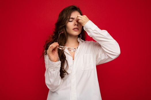 Shot of attractive sorrowful sad upset young curly brunette woman wearing white shirt and optical glasses isolated on red background with free space.
