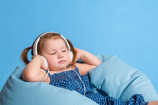 Cute little girl in headphones listening to music using a tablet and smiling while sitting on blue big bag. On blue background.