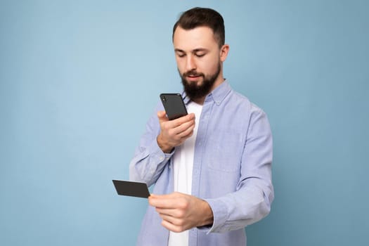 Handsome man wearing everyday clothes isolated on background wall holding and using phone and credit card making payment looking at smartphone screen,