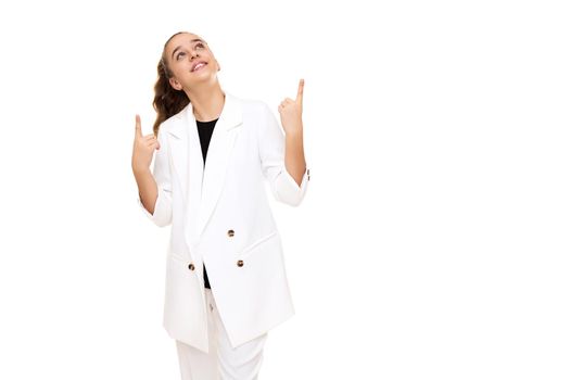 Photo of a beautiful cute adorable smiling brunette teenage girl with a ponytail in a stylish white jacket and white pants isolated on a white background shows a fingers up on a free space for text.