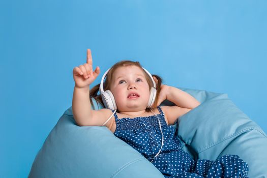 Cute little girl in headphones listening to music using a tablet and smiling while sitting on blue big bag. On blue background.