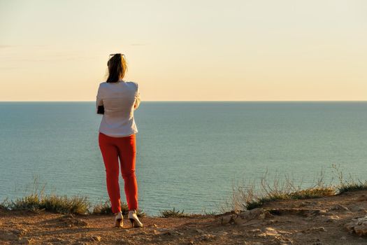 Young beautiful brunete woman in a white jacket and red pants, standing on a cliff over the sea and wait for a call. back view. Copy space. The concept of calmness, silence and unity with nature