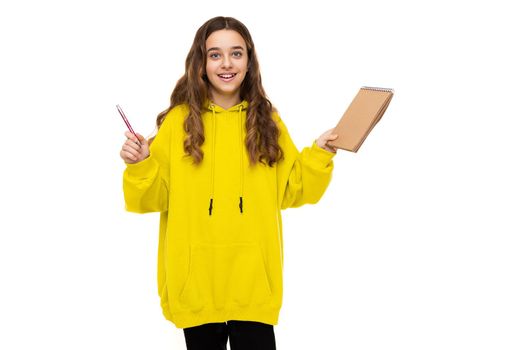 Photo of beautiful adorable cute smiling happy brunette teenage girl in stylish yellow sports hoodie holding brown notebook and pen isolated on white background with copy space for text.